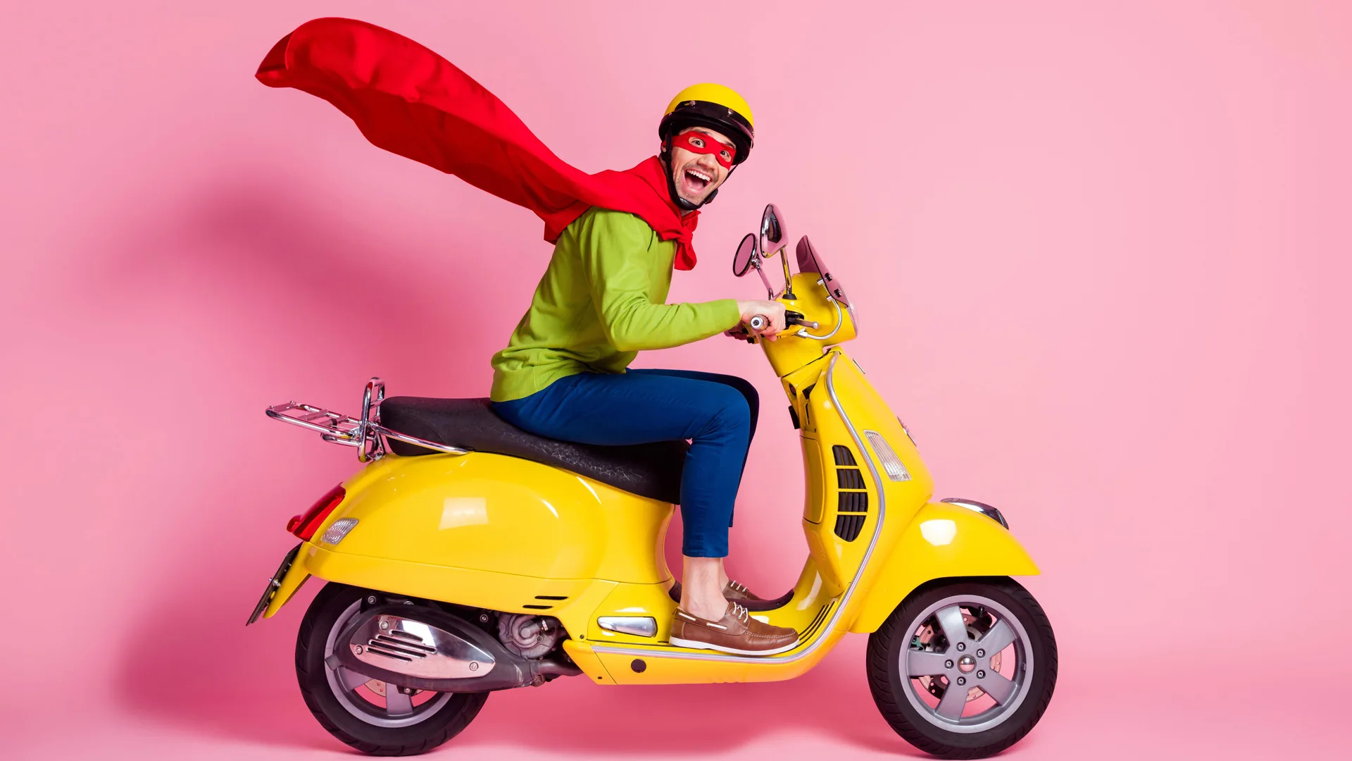 A photograph of a man dressed as a superhero with a red cape and mask riding on a yellow scooter