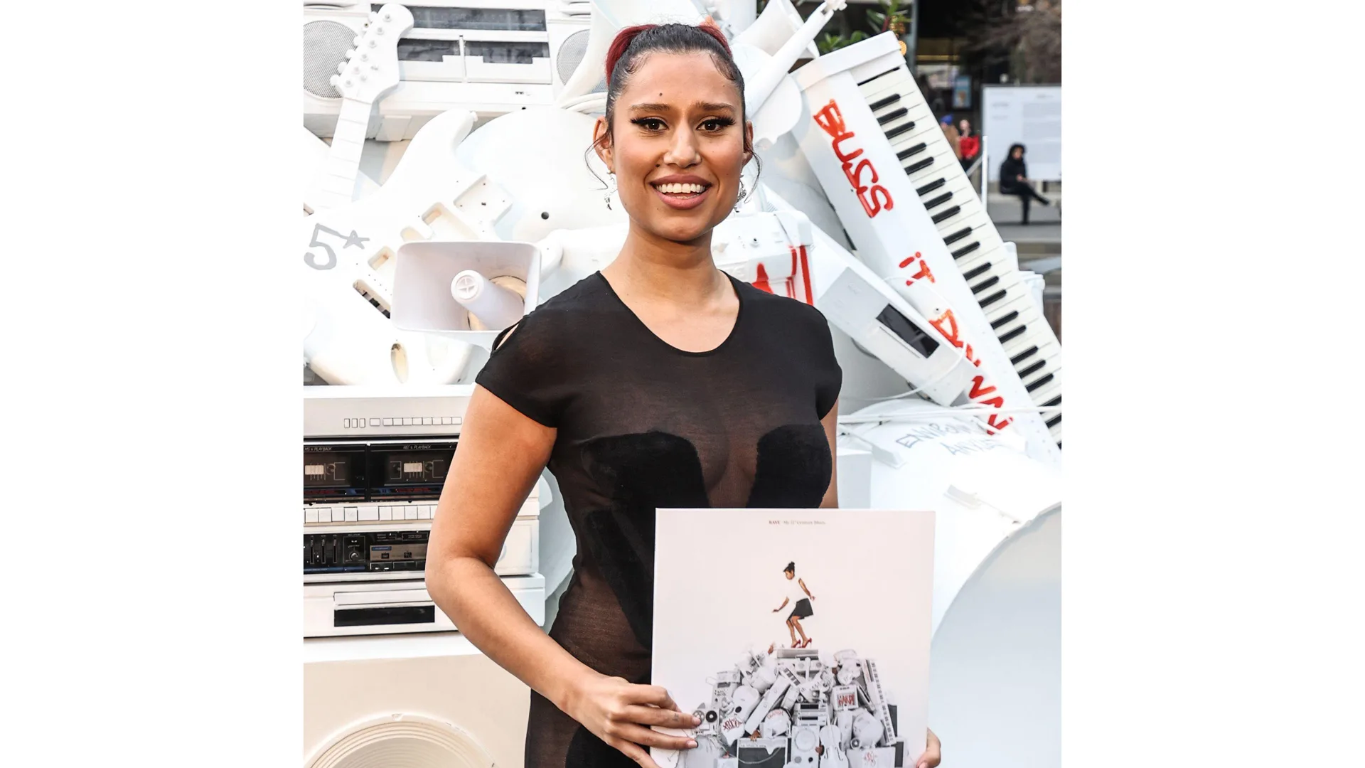 A photograph of the singer Raye holding up her debut album against a background of music related sculptures