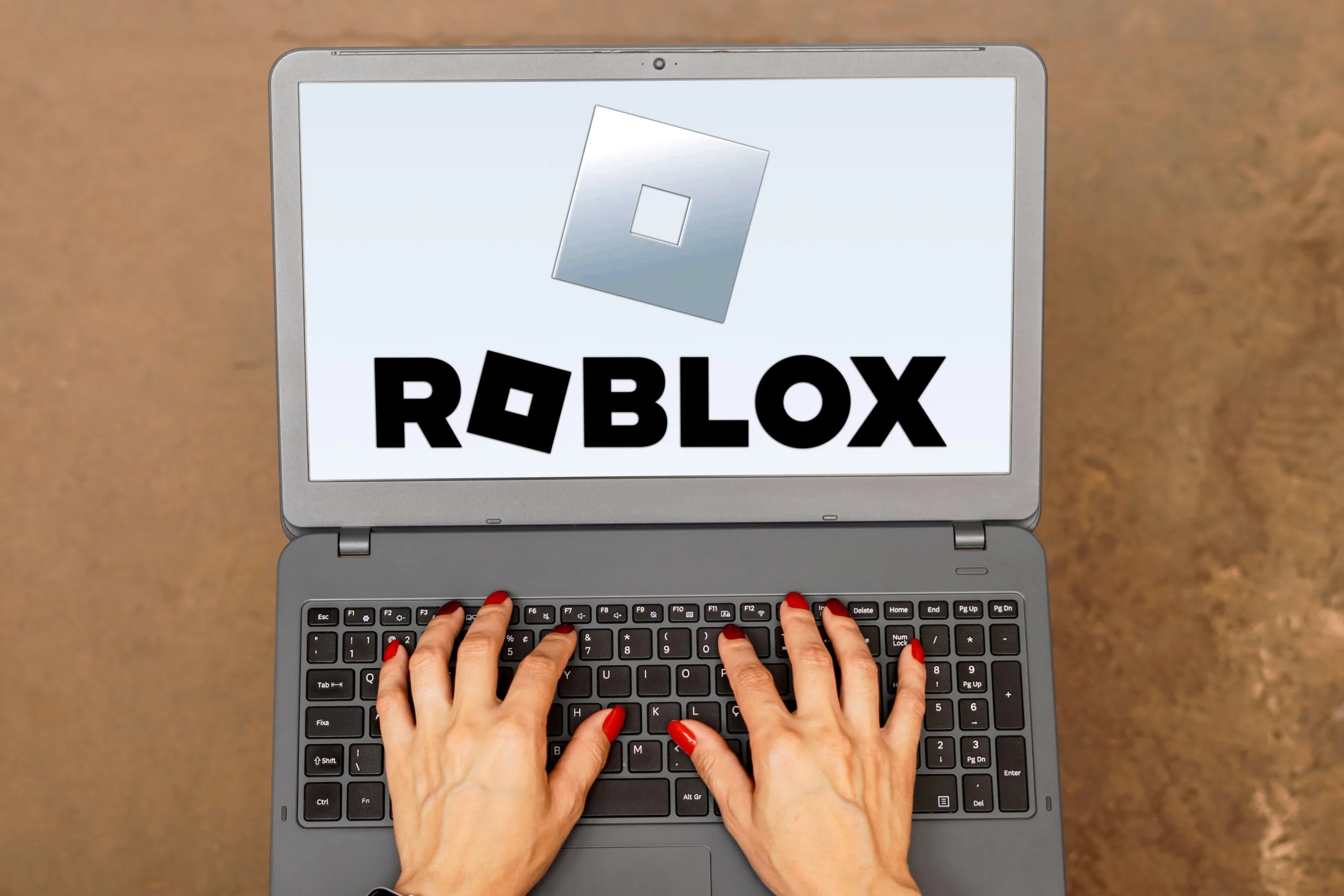A photograph of a pair of hands with red painted nails typing on a laptop with the words ROBLOX on the screen against a beige background