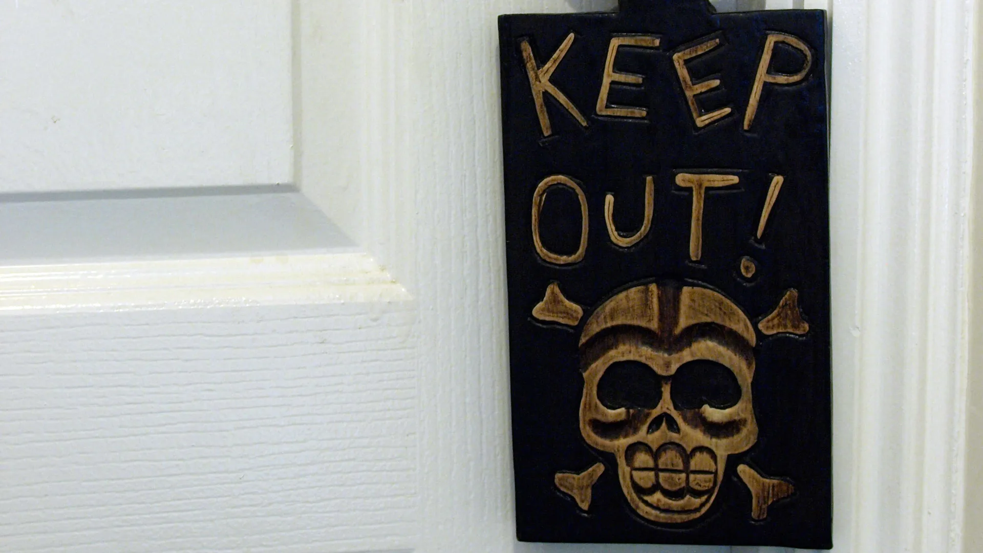 A photograph of a black door sign that says keep out with a gold pirate skull and crossbones hanging on a white door