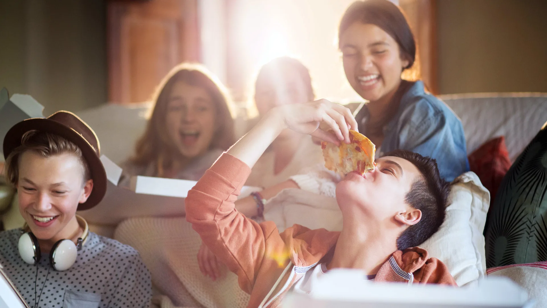 A photograph of some teenagers laughing and eating pizza as the sun shines through the window