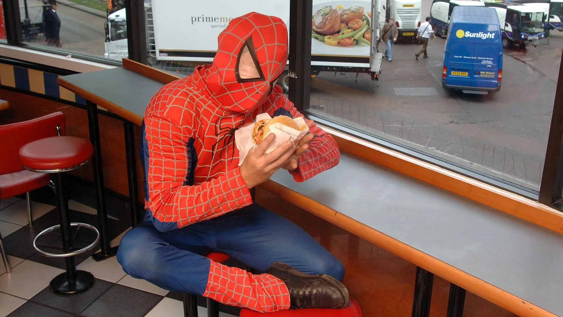 A photograph of a man dressed as Spiderman eating a burger in a restaurant by a window looking out to a street with cars