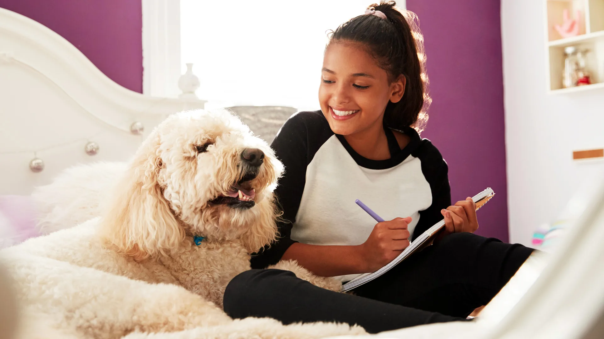 A photograph of a girl sat on her bed doing homework with her pet dog next to her and she is smiling at it