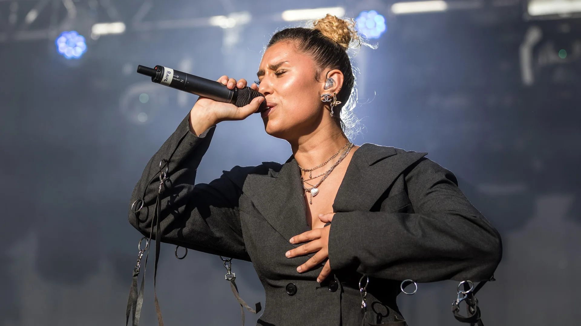 A photograph of the singer Raye singing into a microphone with her eyes closed, wearing a black jacket against a stage background.