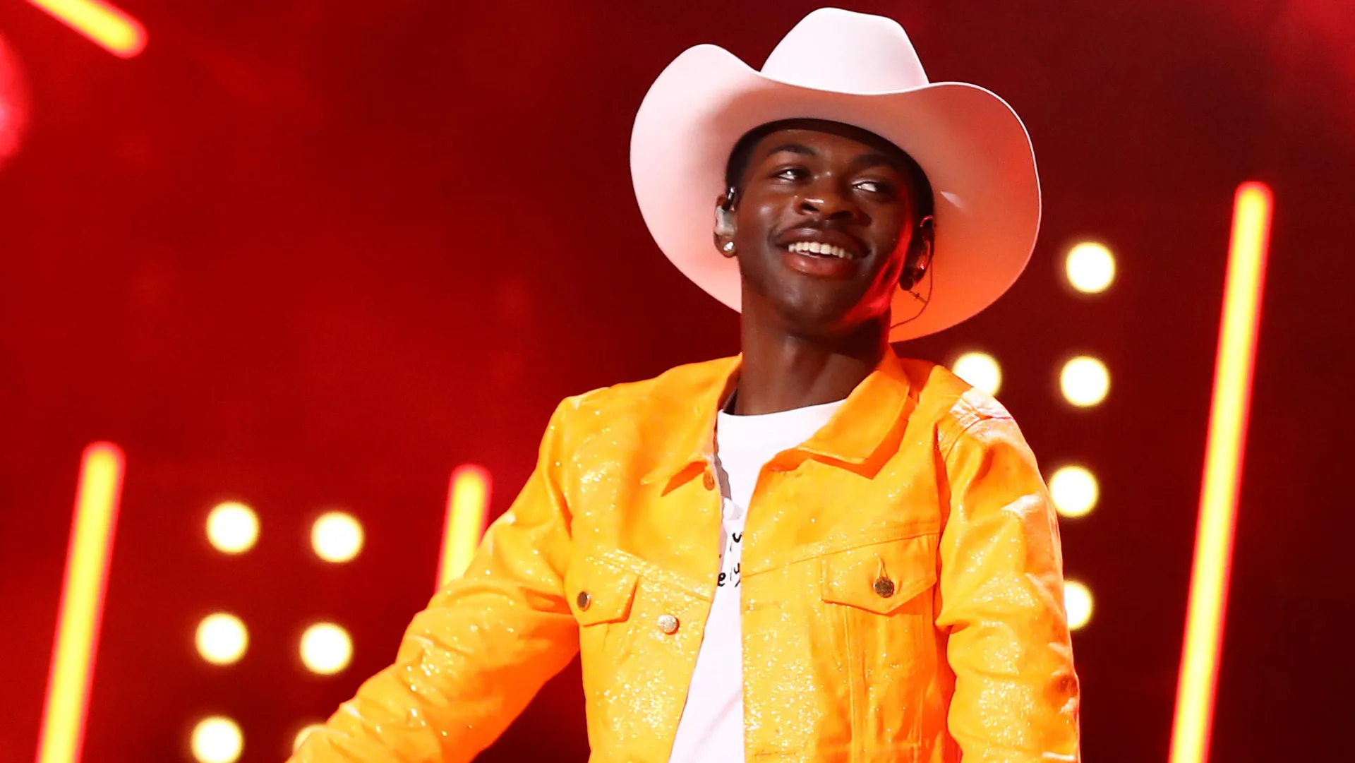 A photograph of Lil Nas wearing a cowboy hat and yellow jacket smiling to the side against a red background with lights