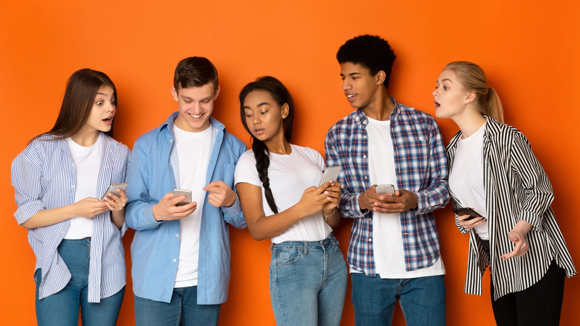 A photograph of teenagers stood in a row on their phones looking at one boys phone. They are all wearing jeans and white tshirts with denim or plaid shirts against an orange background.
