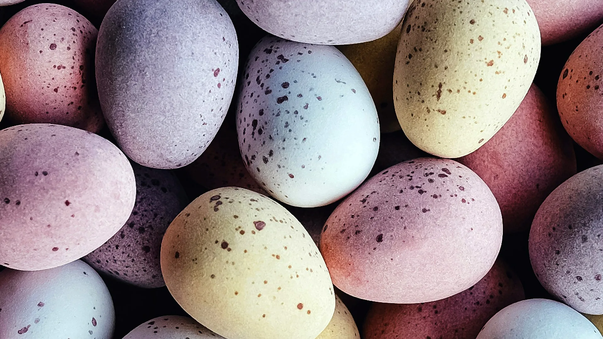 A close-up photograph of speckled chocolate mini eggs in shades of yellow, lilac and pink.