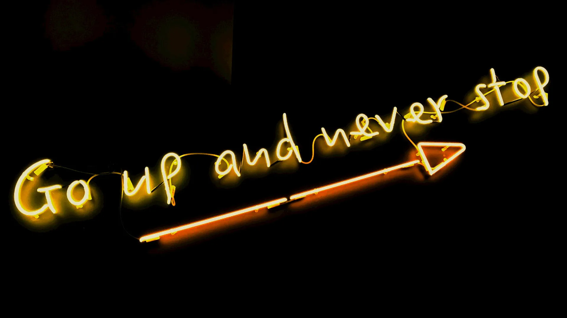 A yellow neon sign that says "Go up and never stop" with an arrow pointing to the right below it against a black background