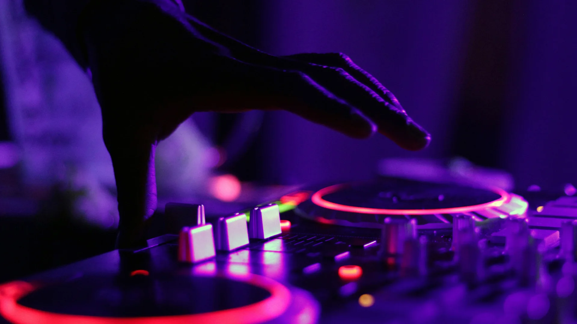 A close up photograph of DJ decks with a hand hovering over to change the buttons lit in neon purples and pinks