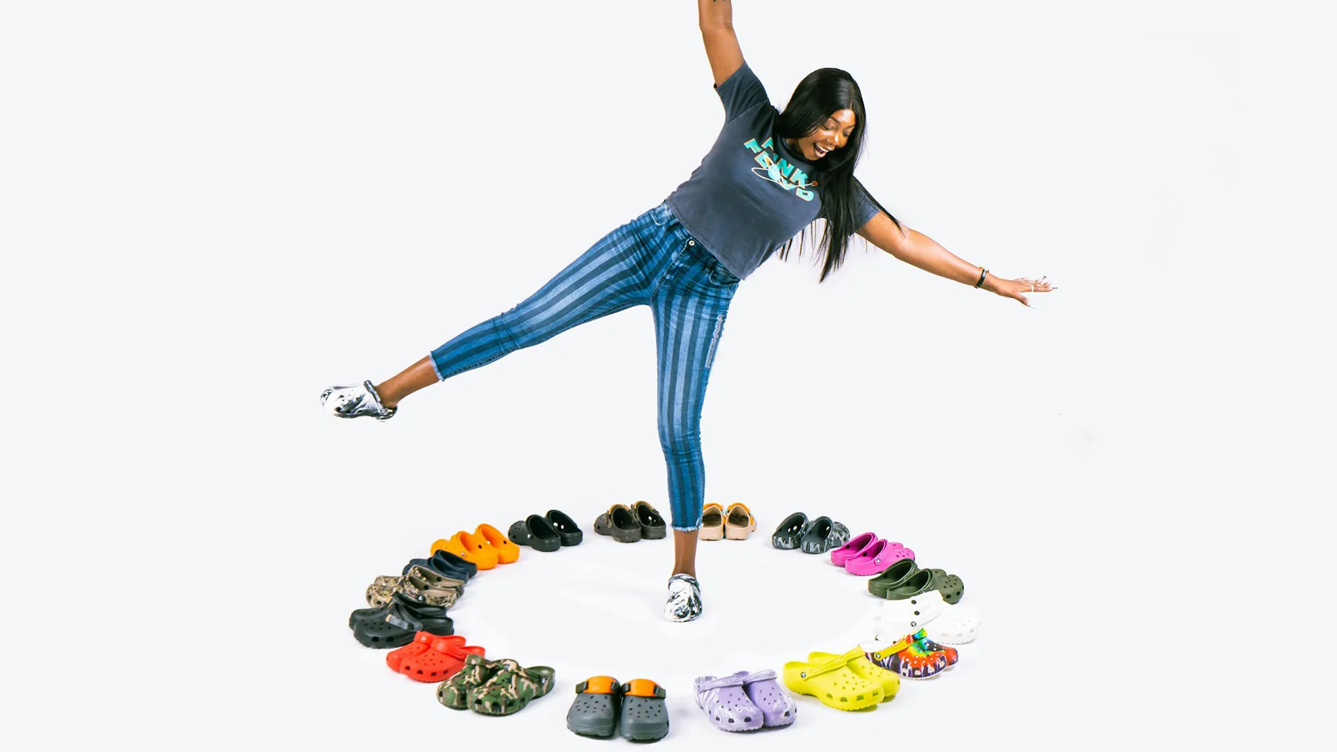Woman standing on one leg in middle of a circle of croc shoes in a white studio 