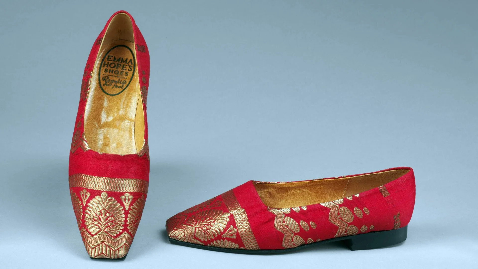 A photograph of some red and gold sari shoes made in 1989 against a grey background