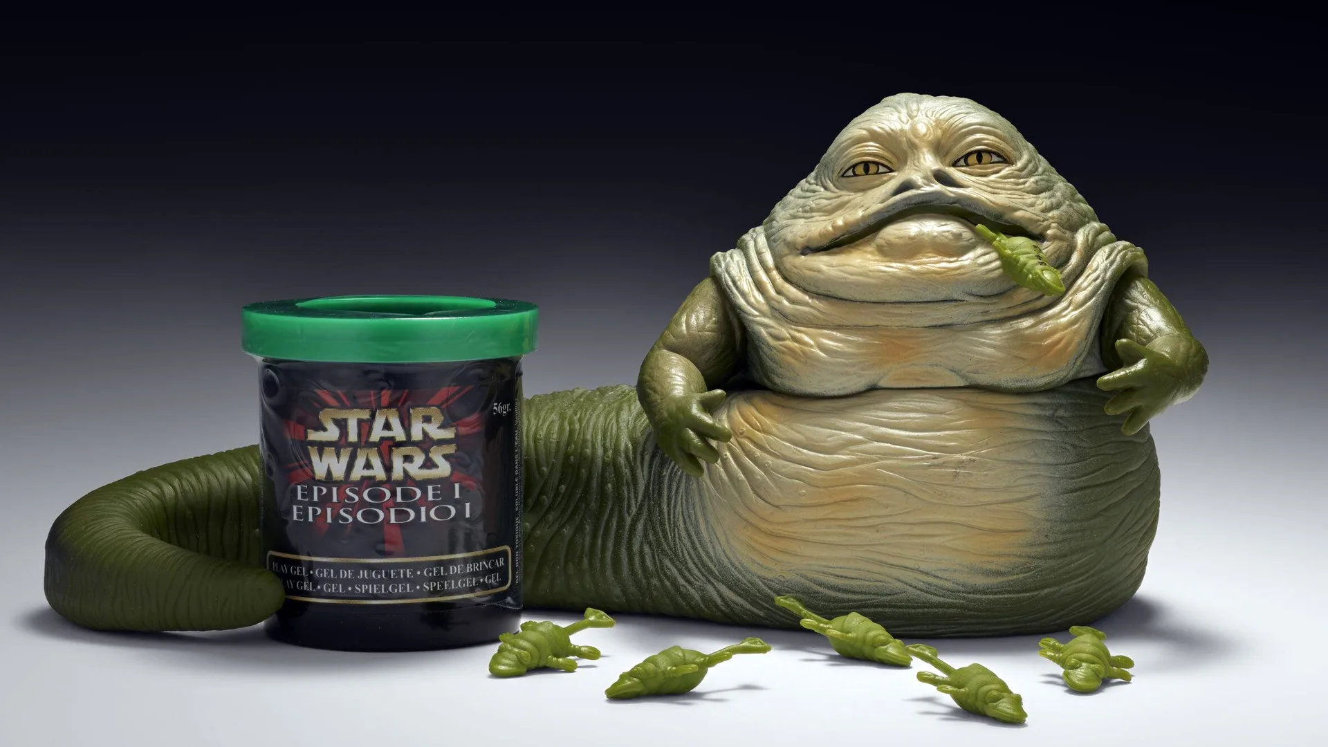 A photo of the V&A collection of a toy Jabba the Glob made by Hasbro for the Star Wars film The Phantom Menace. The toy is green and shows Jabba with a frog hanging out of his mouth next to a tub of Star Wars slime.