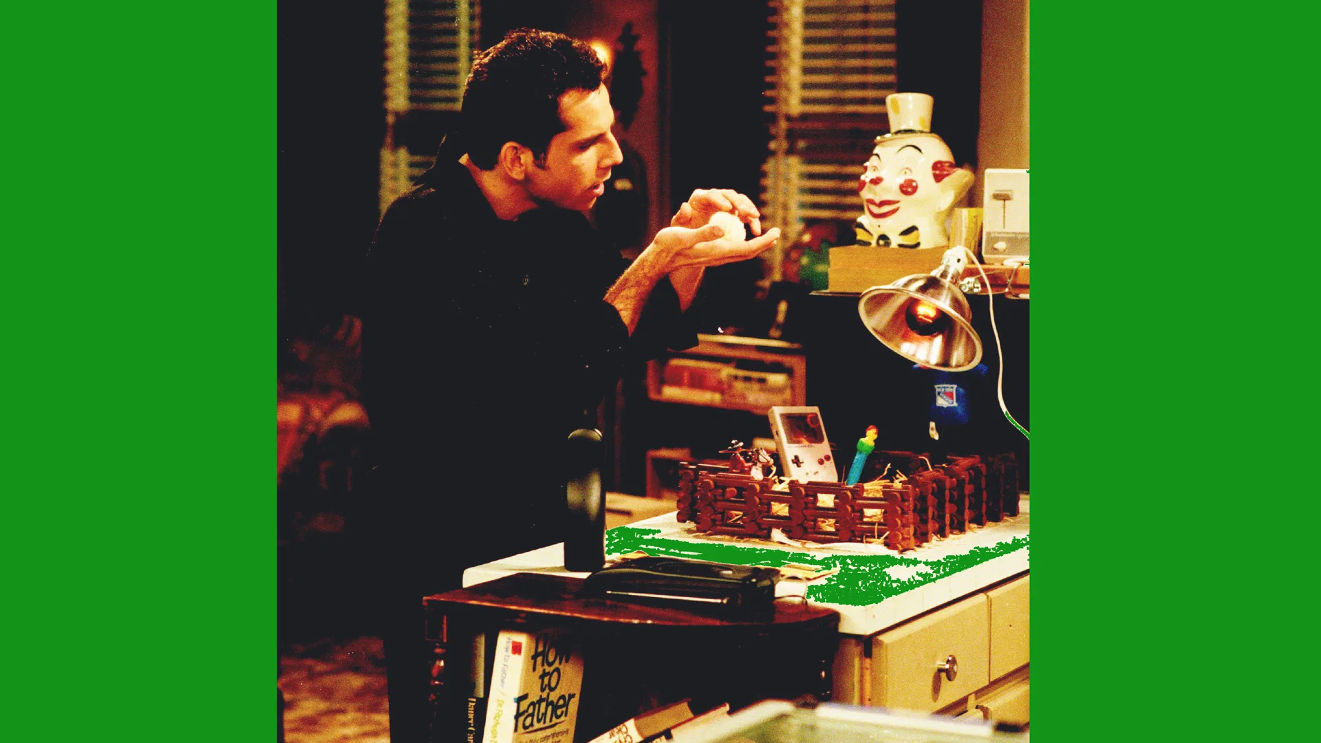 A photograph of the actor Ben Stiller in the series Friends holding a lightbulb next to a lit up clown shaped lamp in a library or livingroom setting