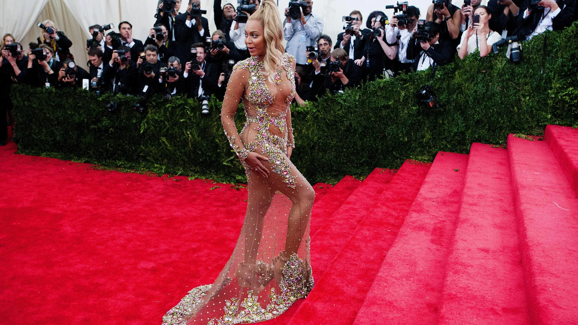 A photograph of Beyonce at the Met Gala in a rhinestone dress with blonde hair in a ponytail - behind her is a group of photographers