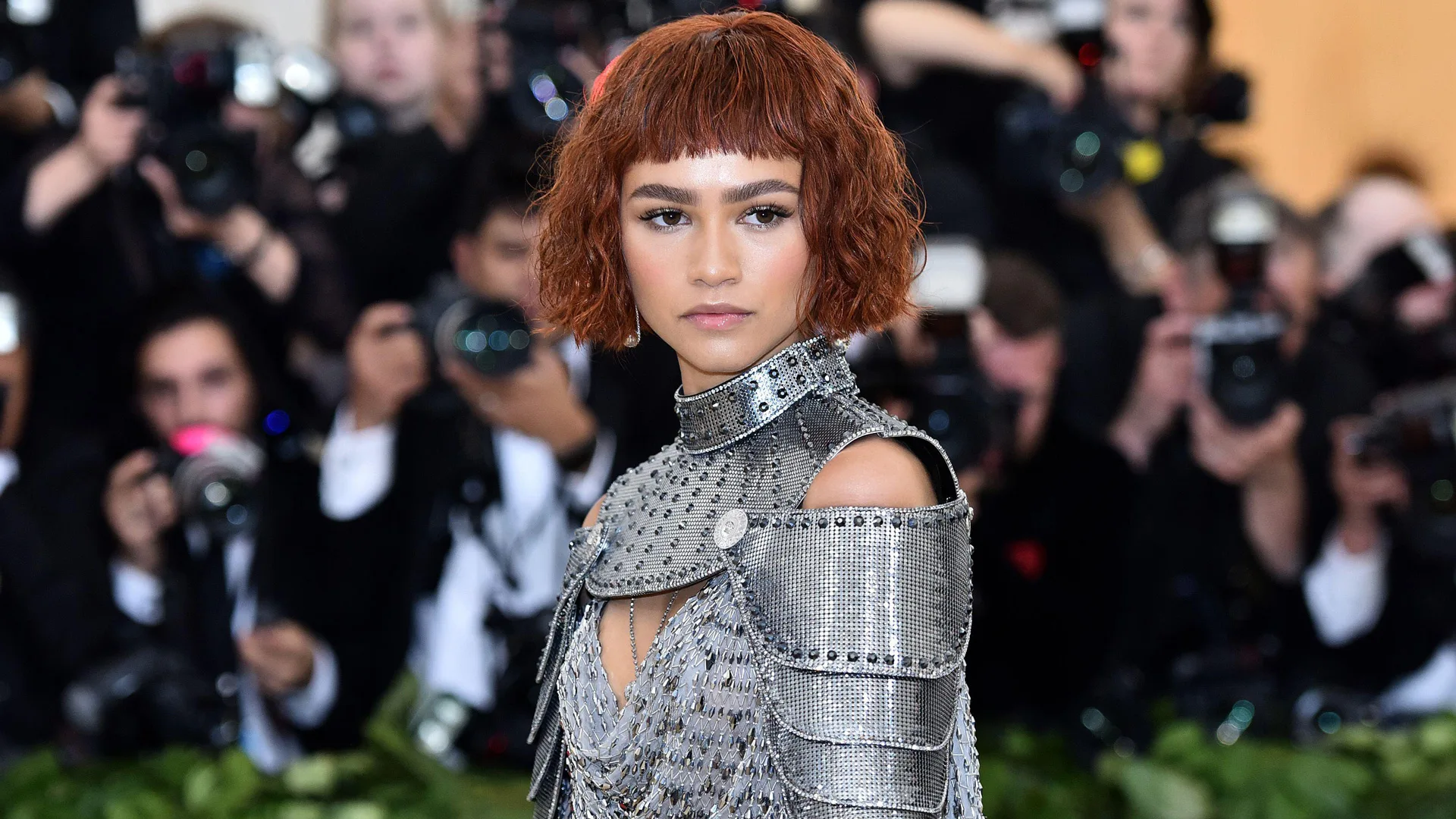 A photo of Zendaya in a suit of armour style outfit with an auburn wig - behind her is a crowd of press photographers
