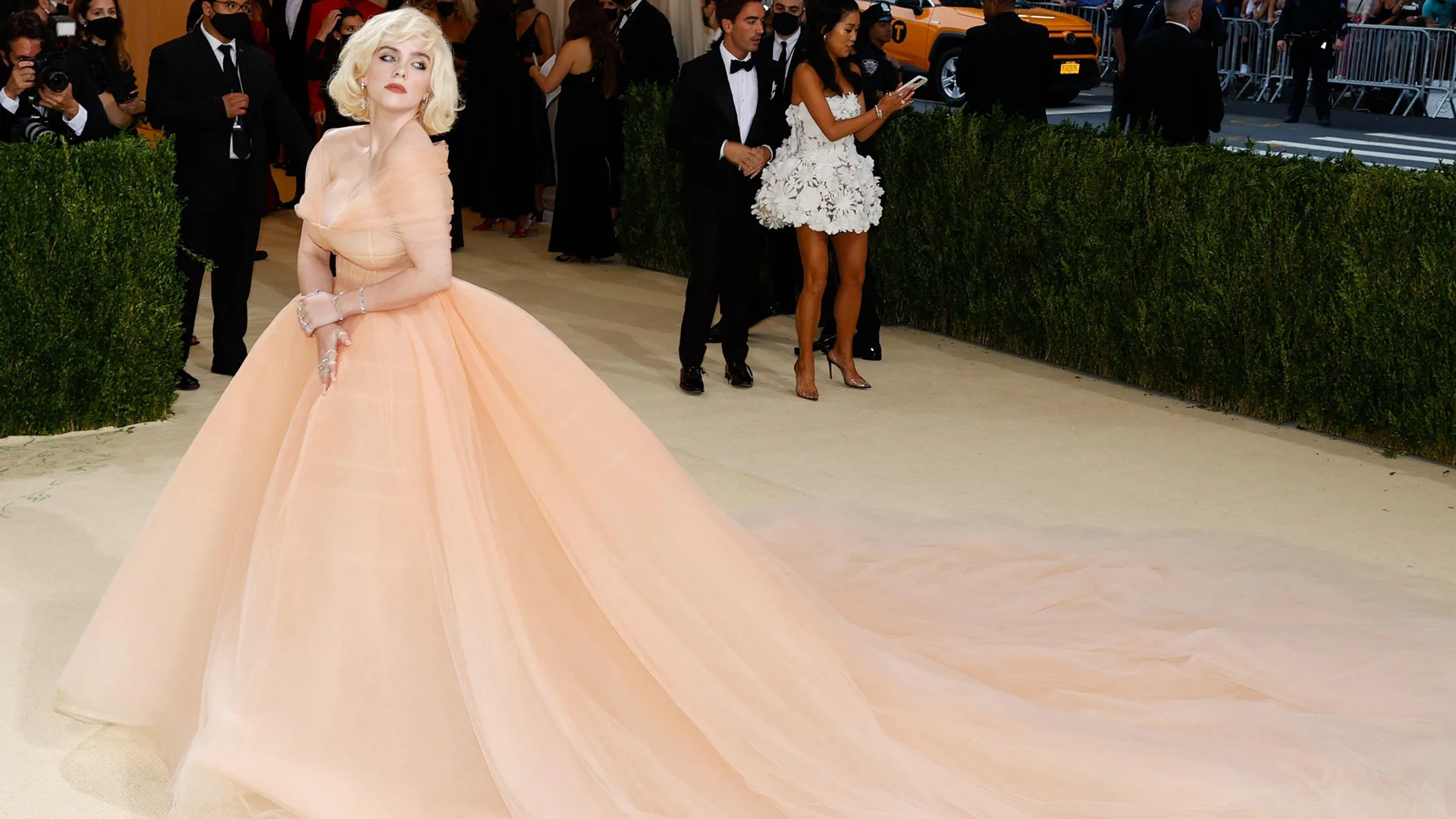 A photograph of Billie Eilish on the carpet at Met Gala wearing a peach coloured ballgown and blonde wig with people in the background on their phones