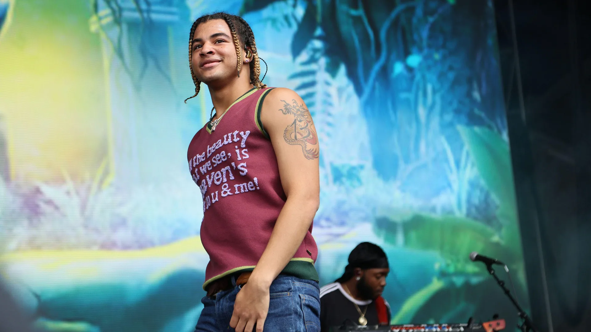 A photograph of the artist 24KGoldn on stage with a red vest against a tropical blue background