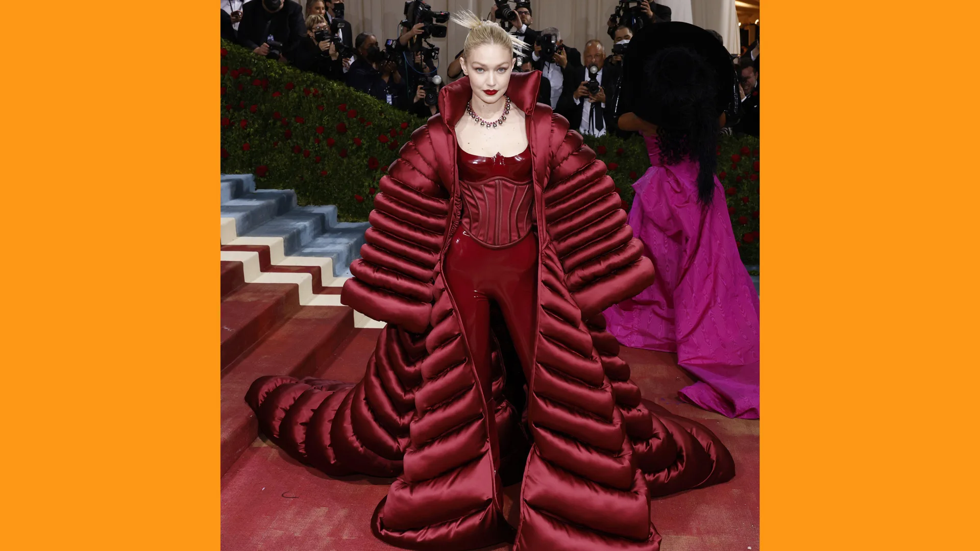 A photograph of Gigi Hadid on the red carpet at Met Gala in a red puffy dress with large sleeves bordered with orange