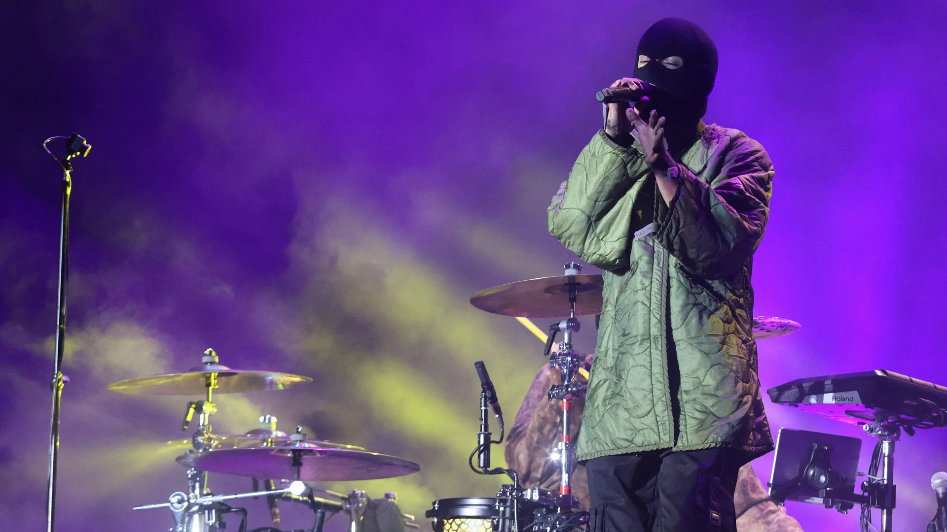 A photograph of the singer of Twenty One Pilots in a black balaclava and green coat on stage with purple and yellow lighting