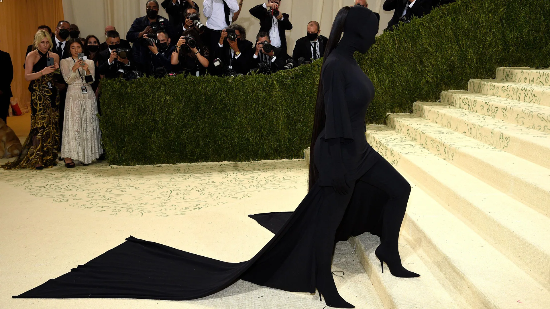 A photo of Kim Kardashian in a full body black outfit by Balengia at the Met Gala on the steps walking up with the press taking photos behind her