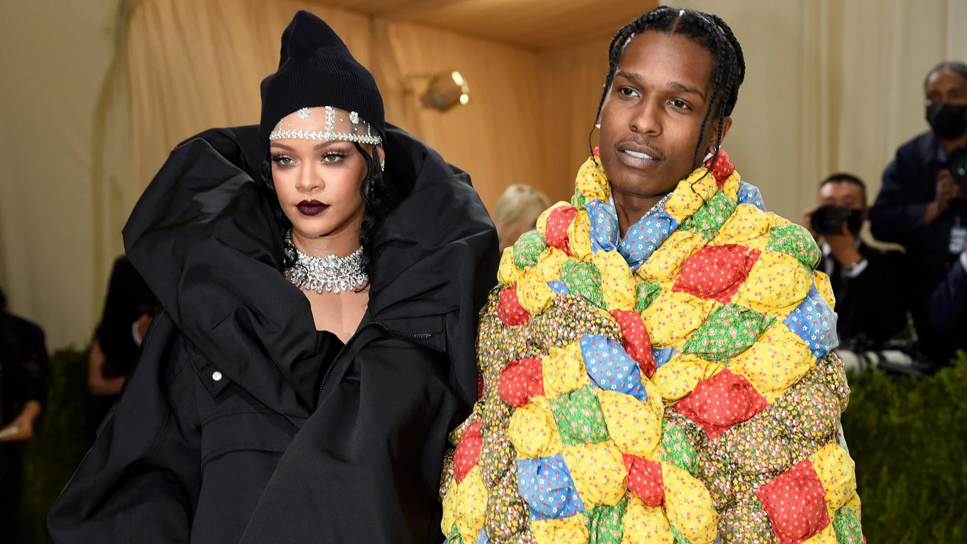 A photograph of Rhianna and ASAP Rocky at the Met Gala. Rhianna is wearing a black ruff neck outfit and hat with jewellery and ASAP Rocky is wearing a multi-coloured blanket style cape