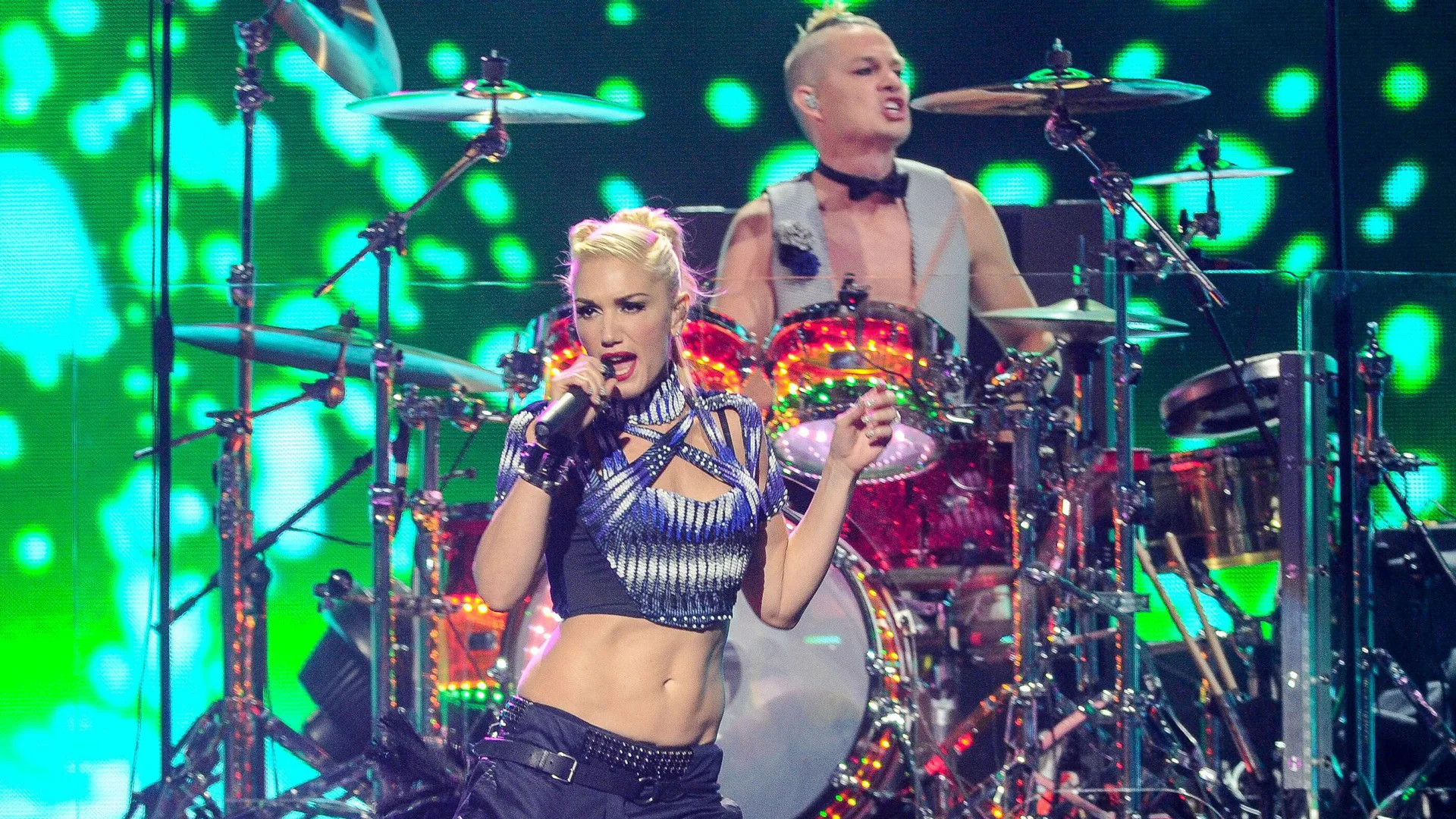 A photo of Gwen Stefani performing on stage in a cropped top with her drummer behind against green lights