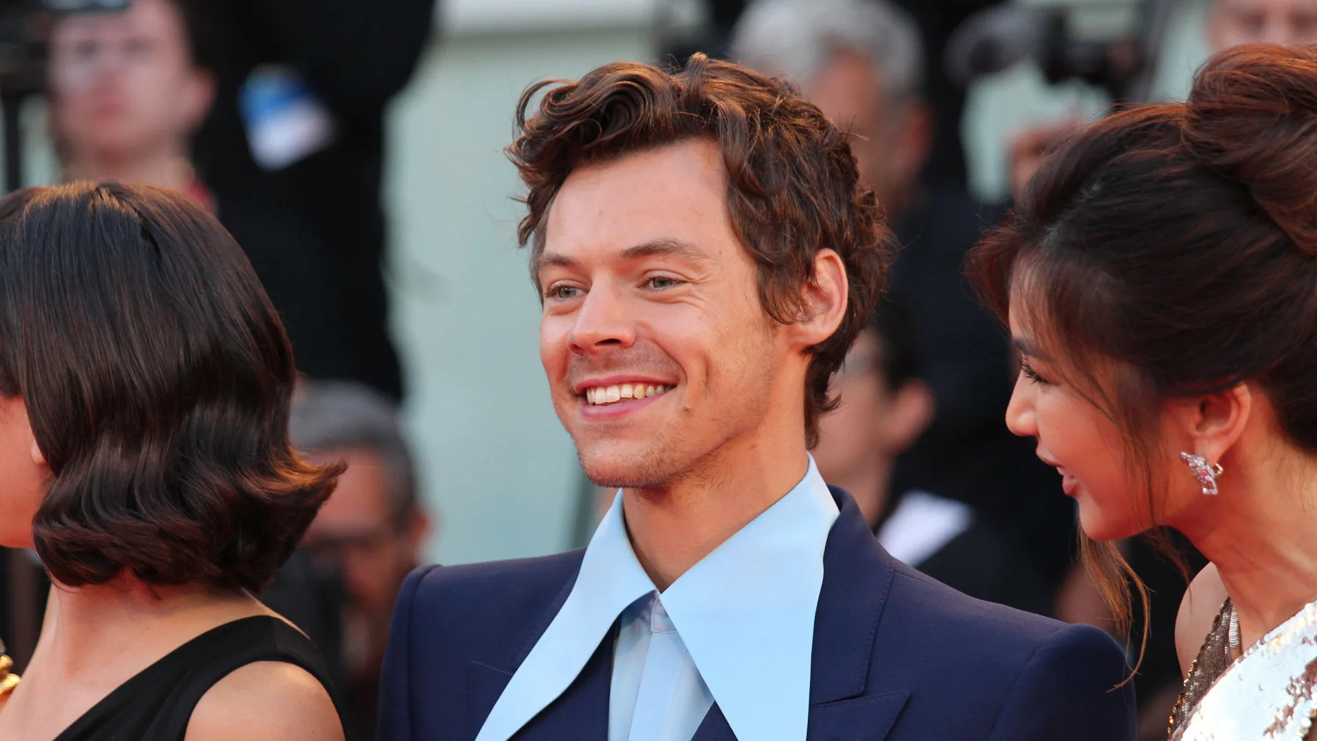 A photograph of Harry Styles wearing a big blue collared shirt smiling to someone off camera
