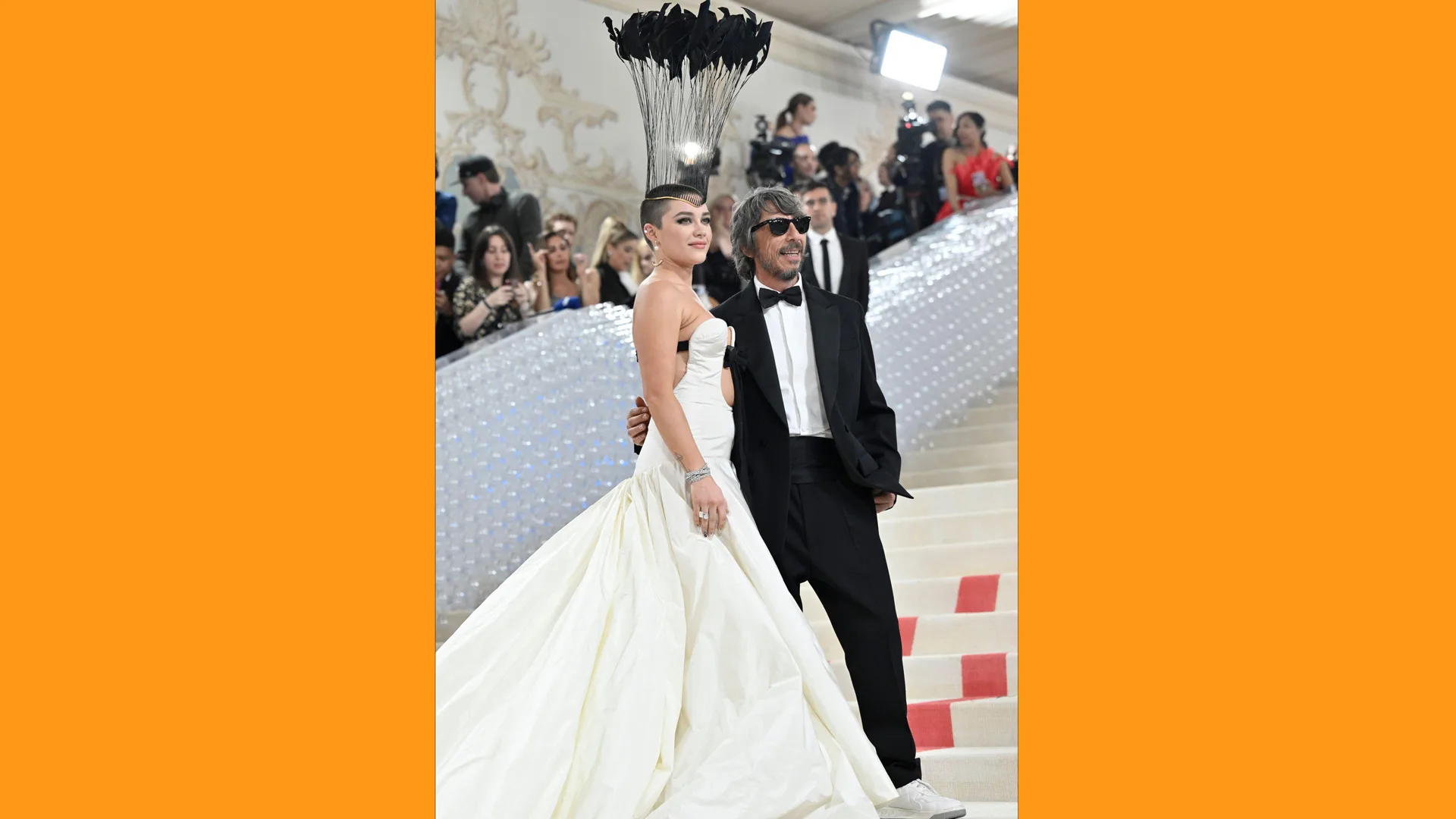 A photograph of Florence Pugh in a white gown with a black feather hat with a man in a black and white suit on the carpet of the Met Gala with orange borders