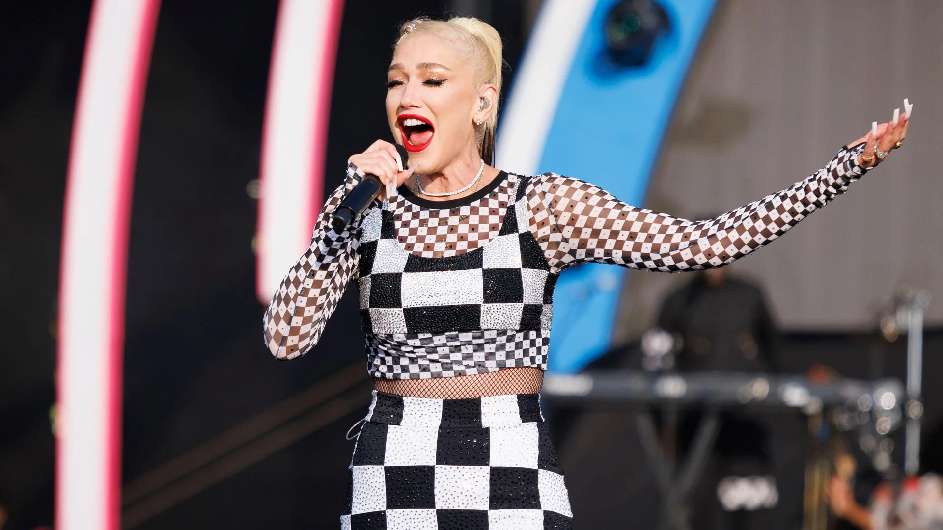 A photograph of Gwen Stefani singing on stage in a black and white outfit against a blue and red lit background