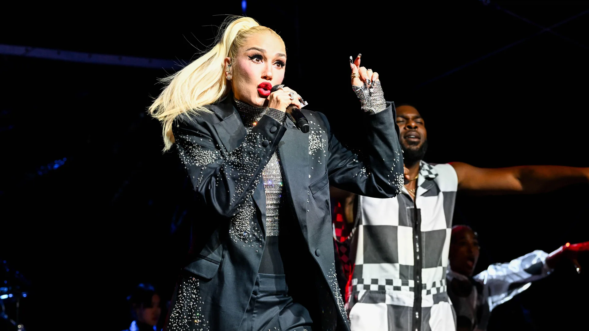 A photograph of Gwen Stefani performing on stage in a black outfits with her band member in a black and white outfit next to her