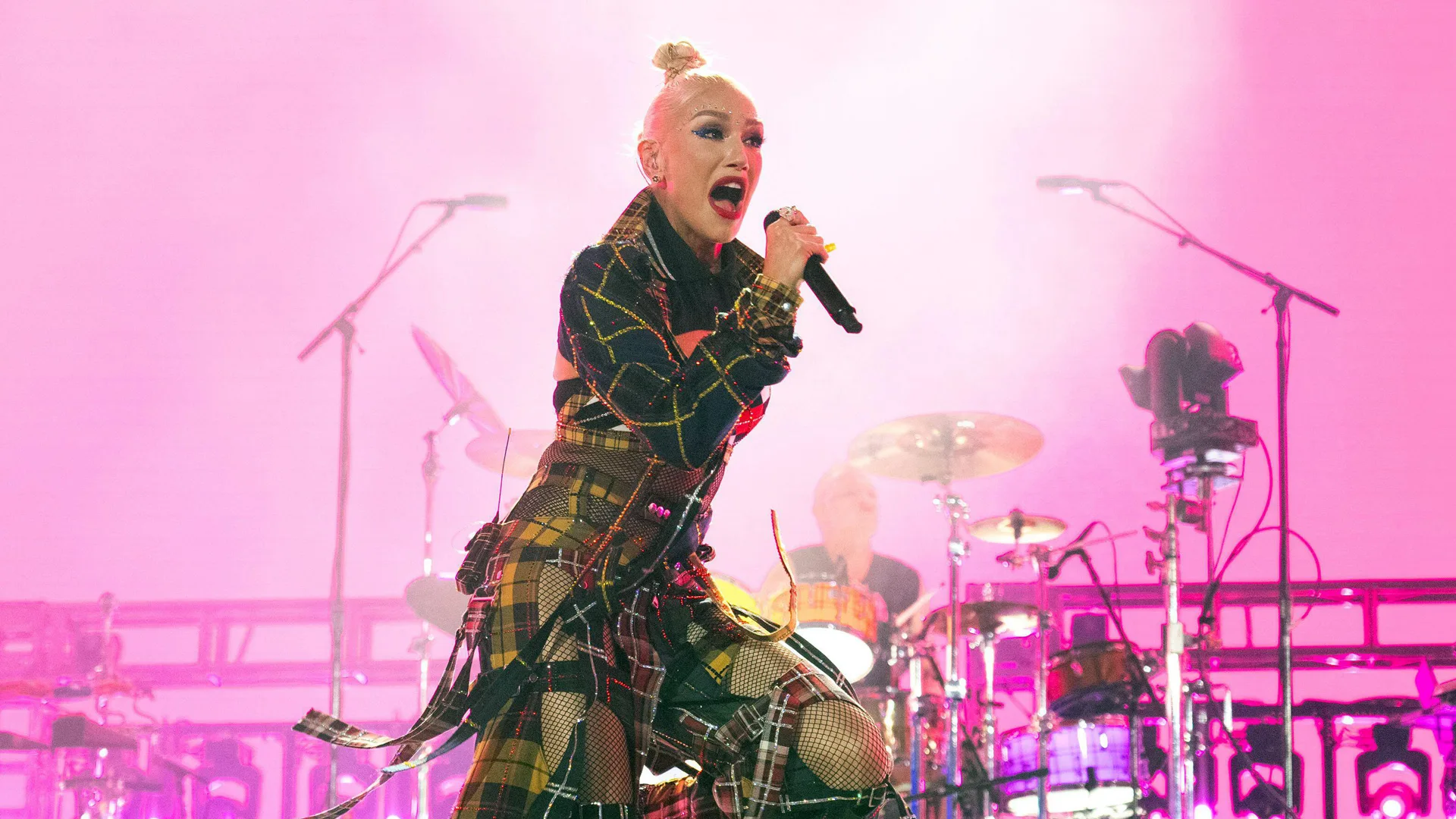 A photo of Gwen Stefani performing on stage singing into a mic against a pink lit background