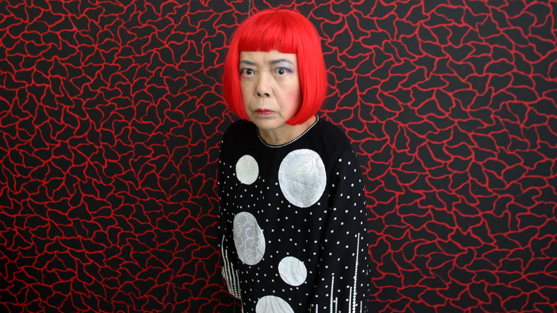 A photograph of Yayoi Kusama with her red wig and black and white spotted top against a red and black lined odd shape background