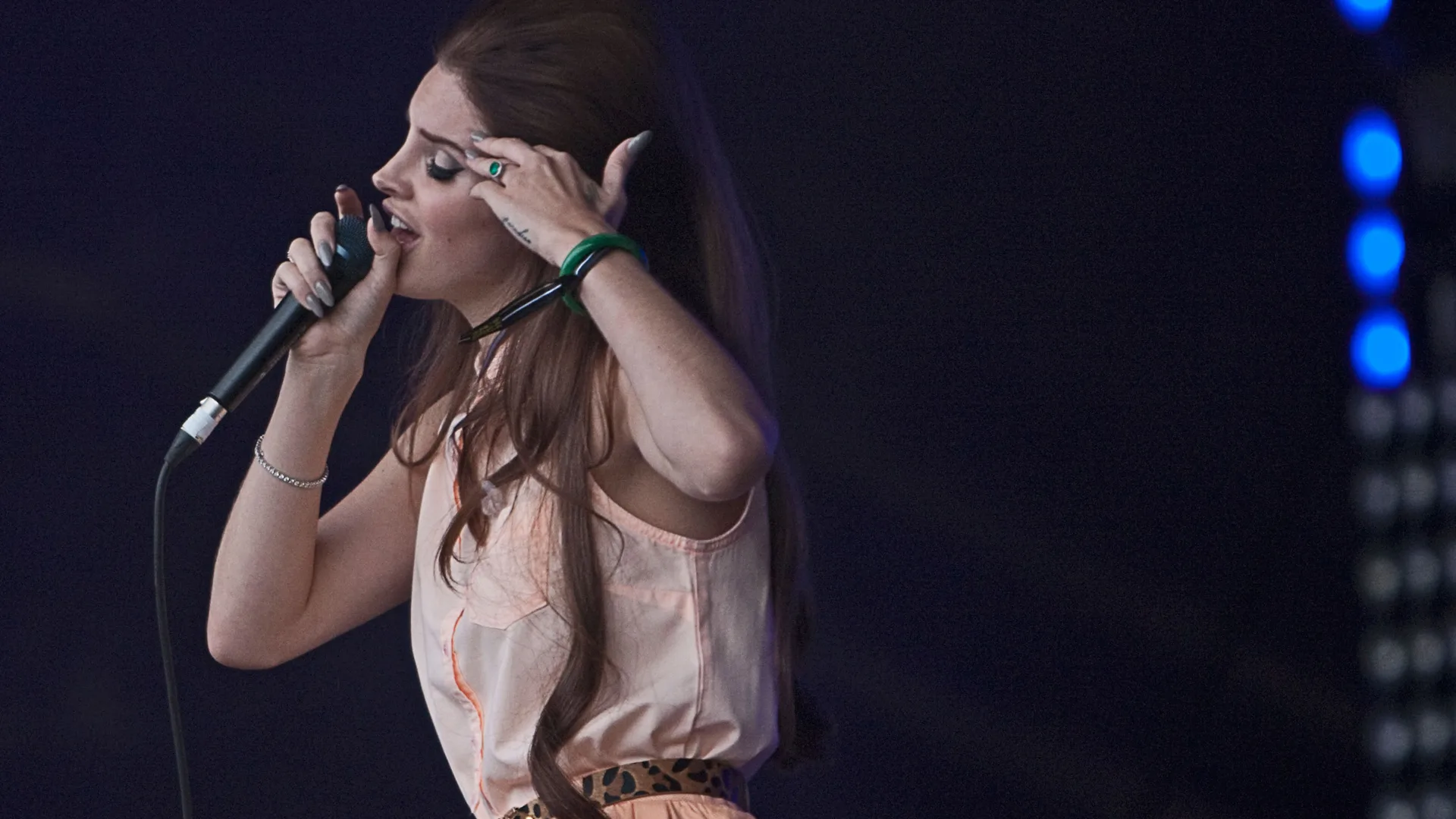 A photograph of Lana Del Ray from the side singing on stage in a pink sleeveless shirt