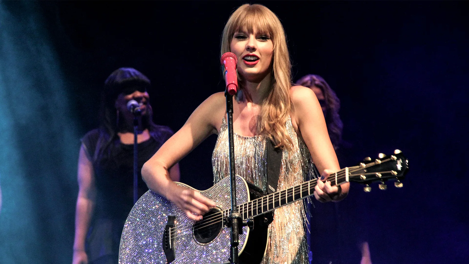 Taylor Swift with acoustic  sparkly silver guitar wearing a silver dress in front of some backing singers 