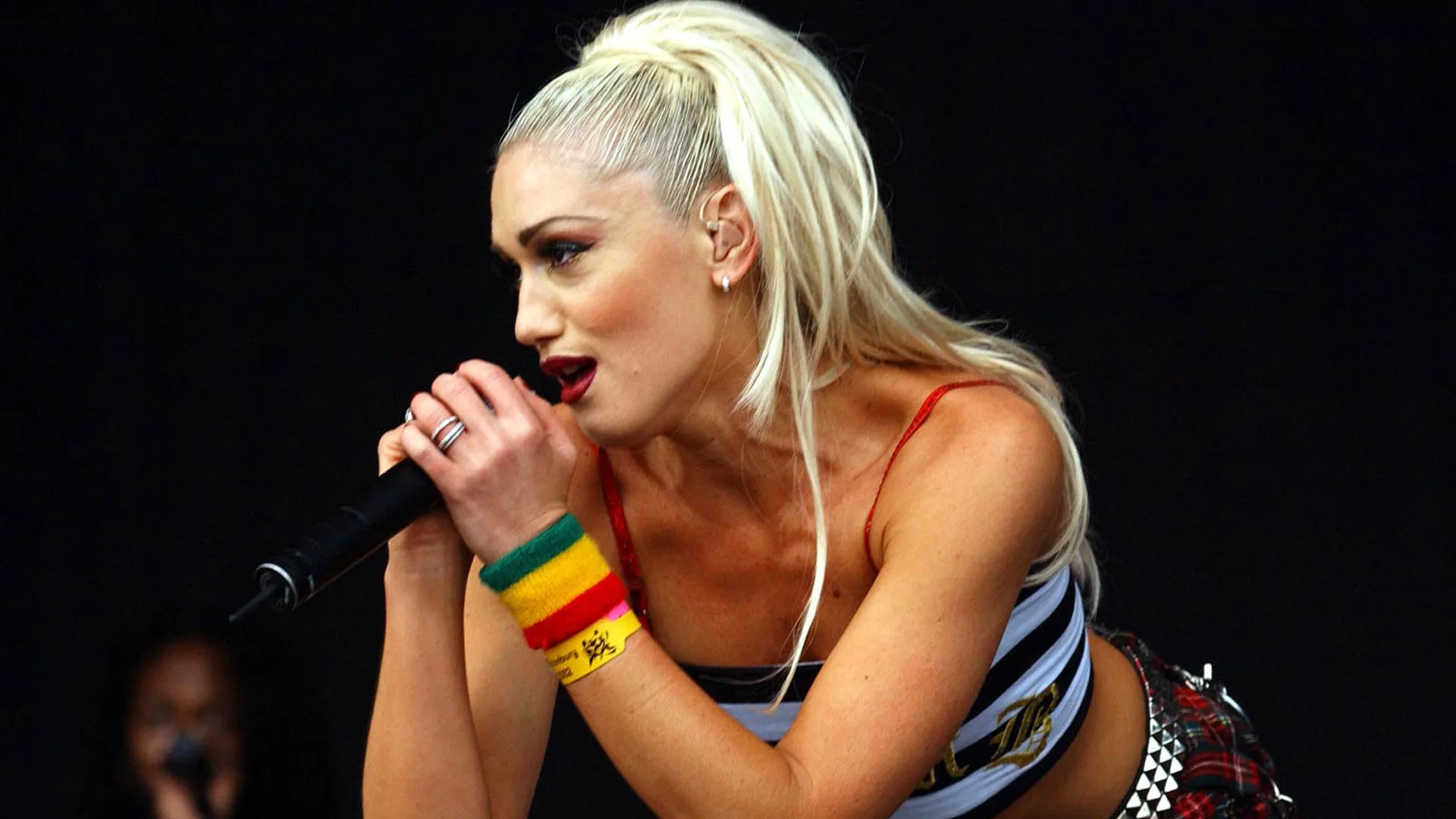 A photograph of Gwen Stefani singing into a mic wearing colourful bands on her wrist and a striped top.