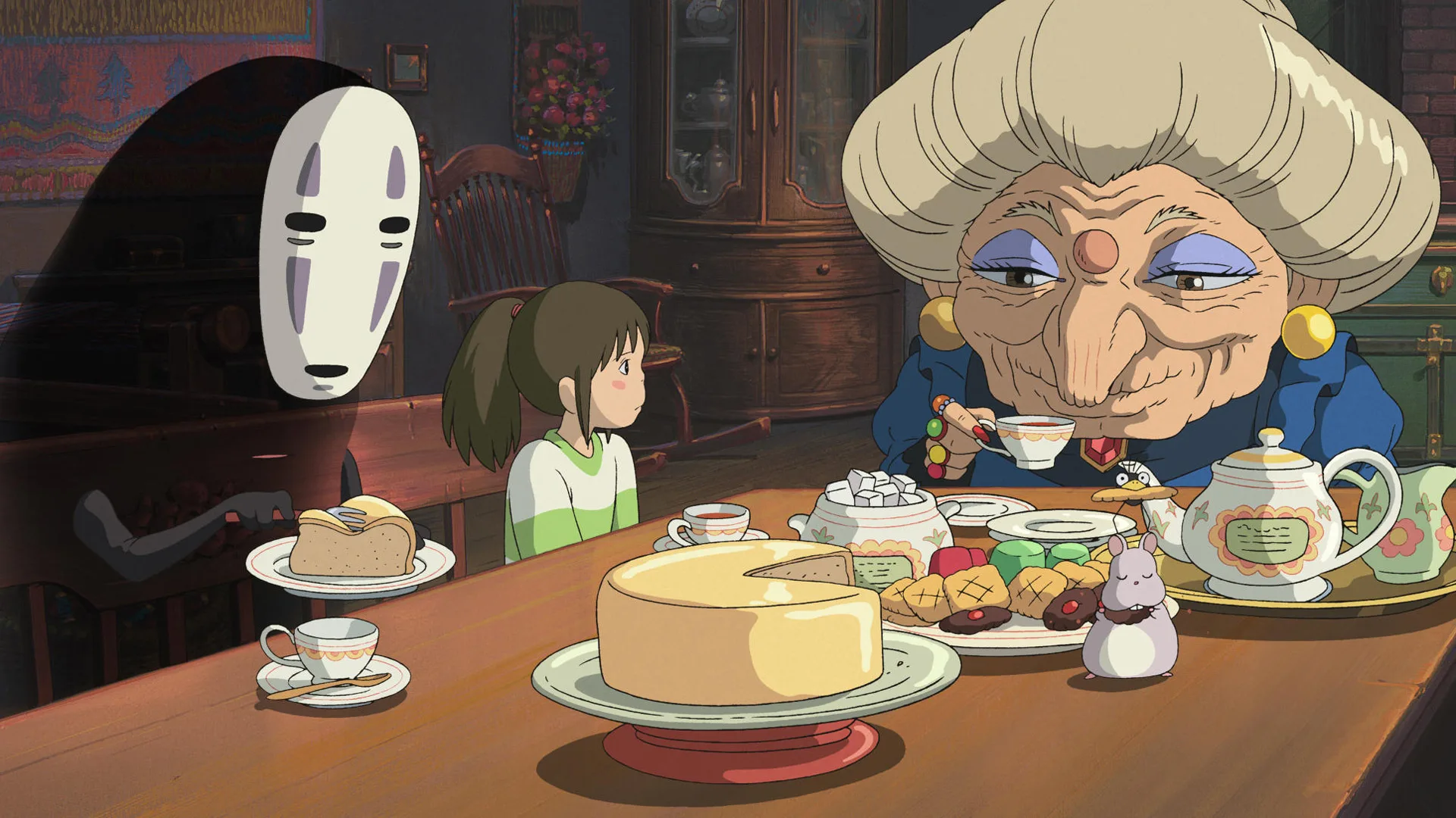 A still from the animated film Spirited Away showing the characters Zenith, No-Face and Chihiro at a table full of food