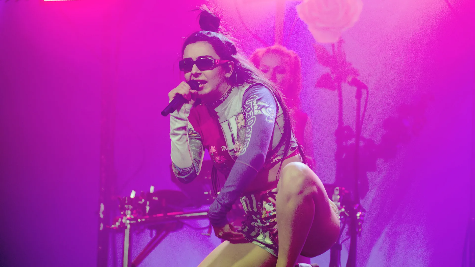 A photograph of the singer Charli Xcx on stage with a mic and sunglasses crouching down against a background of pink lights
