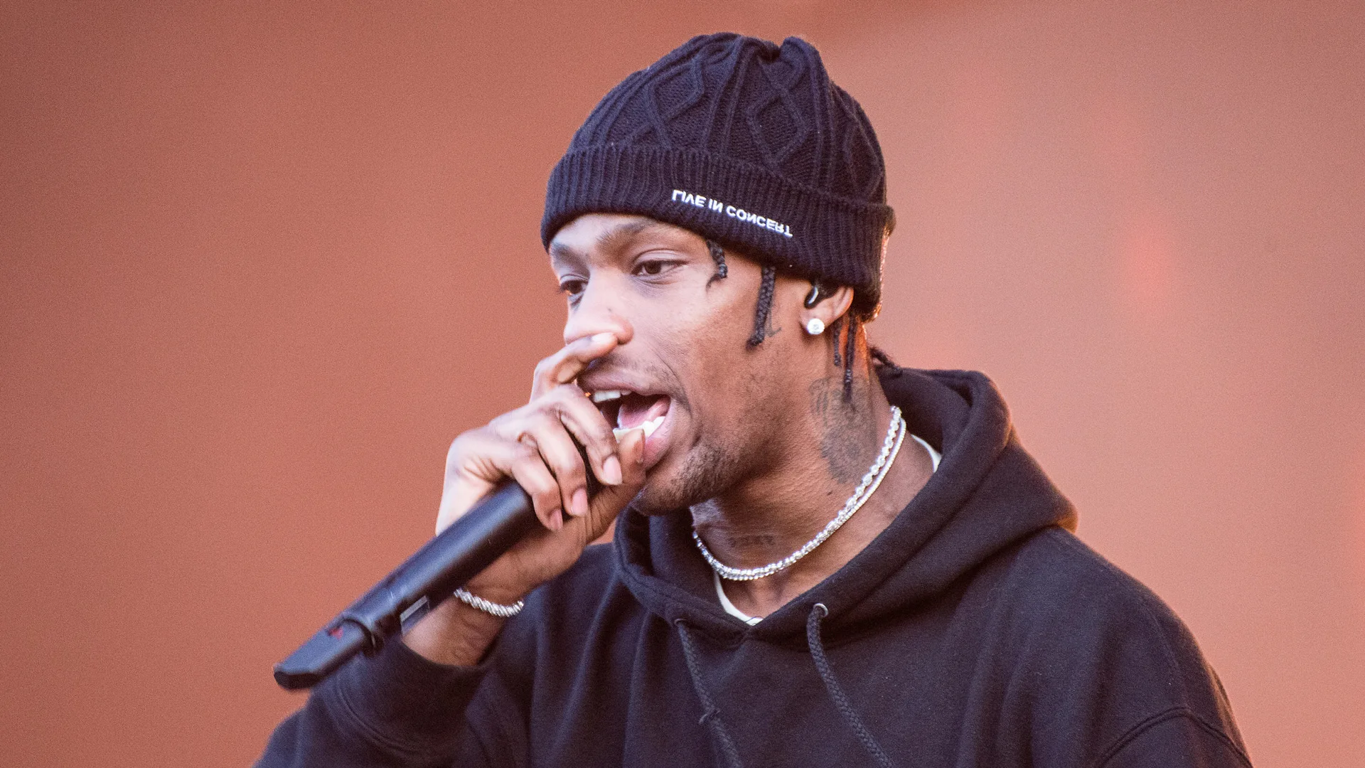 A photograph of Travis Scott singing into a microphone wearing a black beanie hat and hoodie against a terracotta background