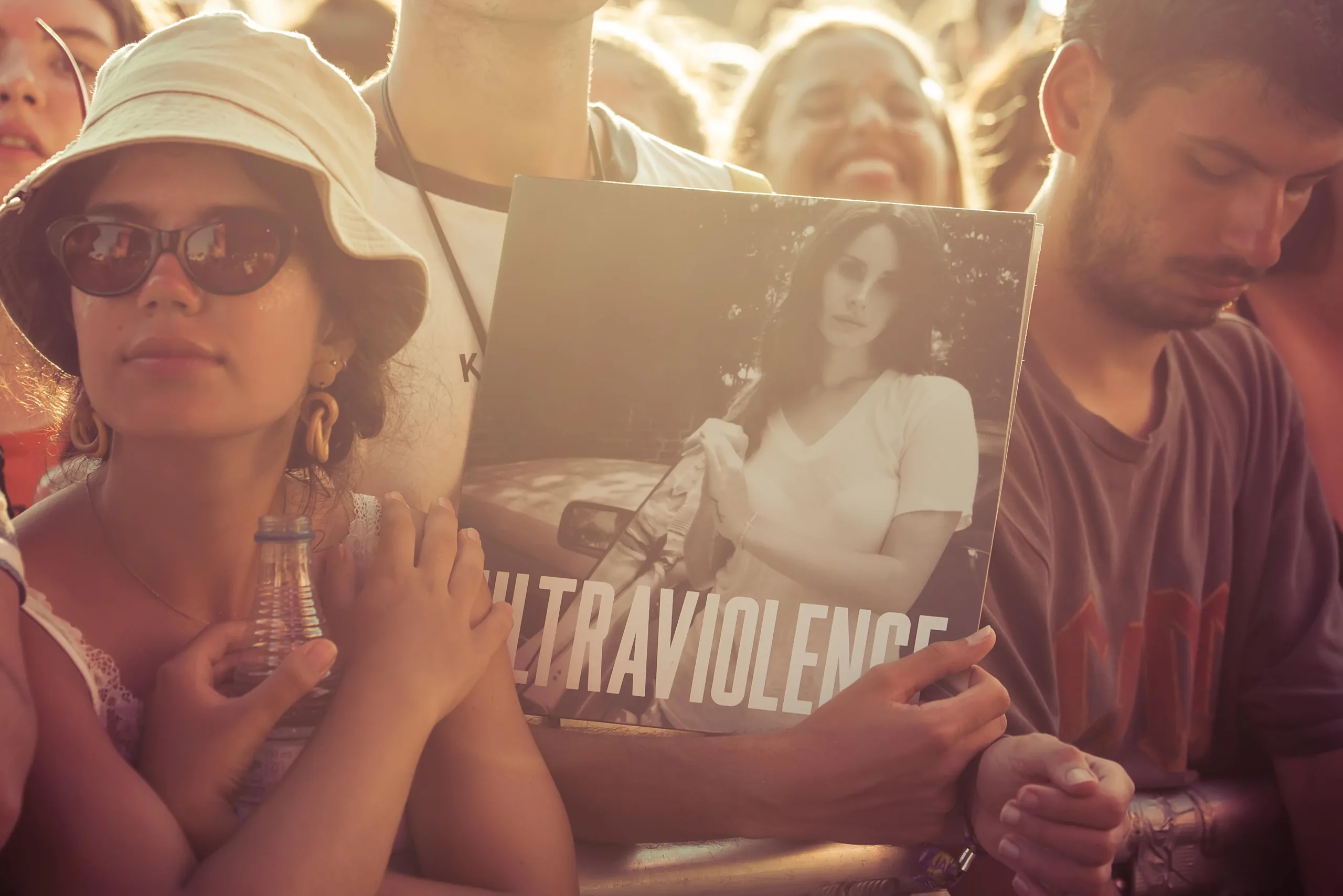A photograph of someone holding up Lana Del Ray's album Ultraviolence next to a girl in sunglasses and a white bucket hat with the sun shining on them