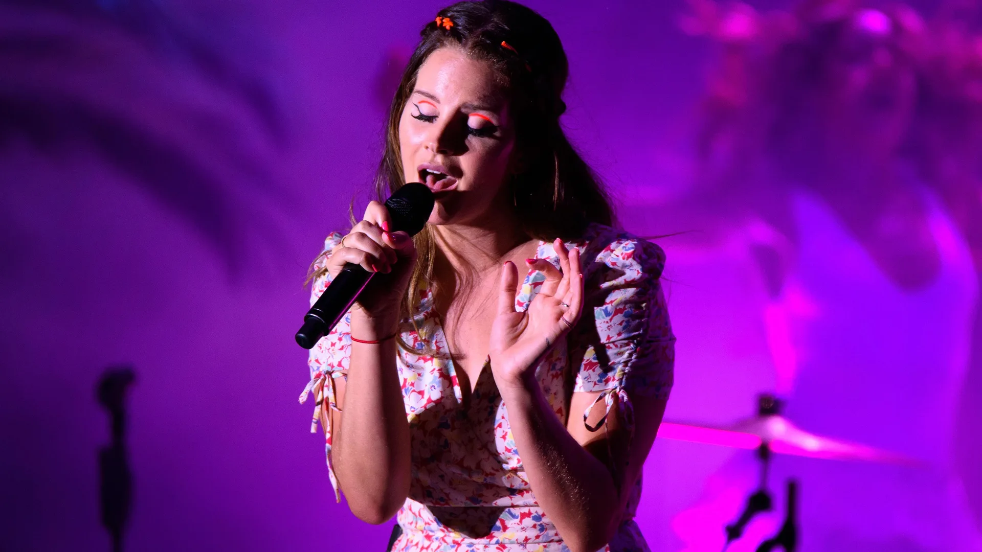 A photograph of Lana Del Ray singing on stage in a floral dress against a purple pink lit background