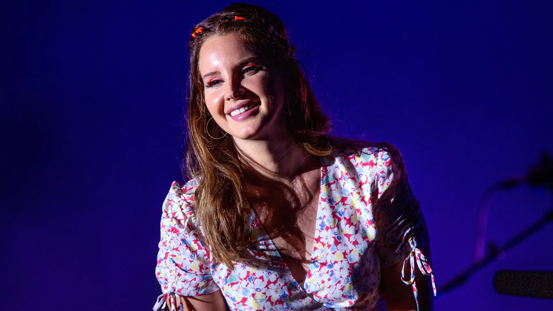 A photograph of Lana Del Ray smiling off camera wearing a floral dress against a dark blue background