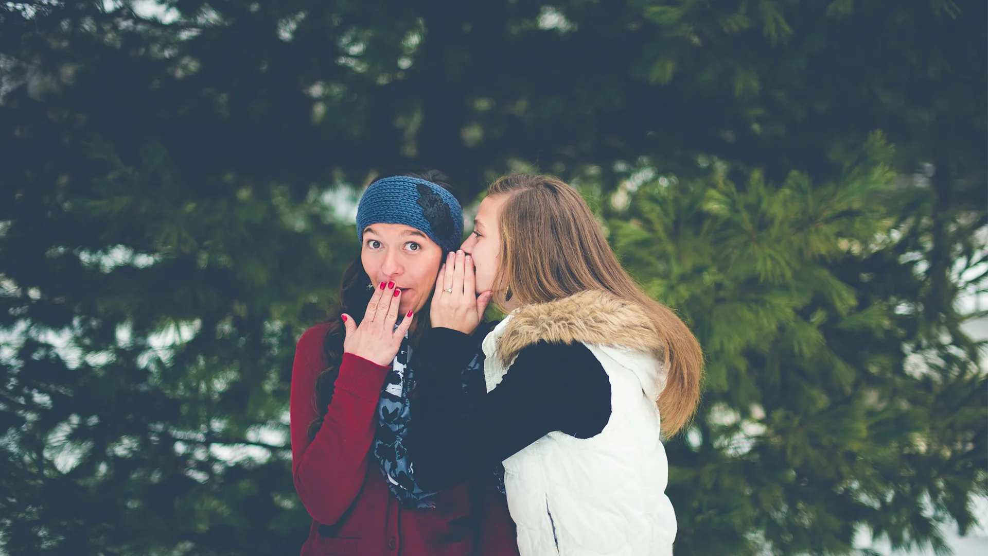 Woman whispering into another womans ear as they stand infront of a tree