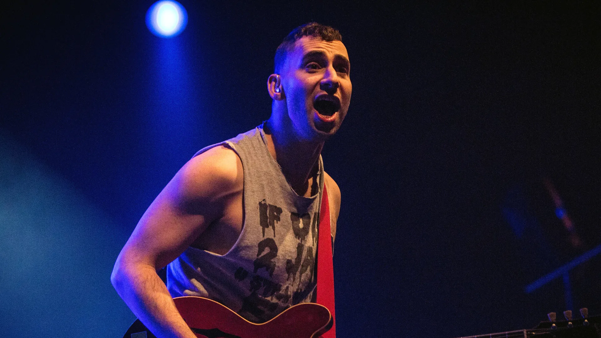 A photograph of the lead singer of The Bleachers performing on stage with a purple light shining on his face.