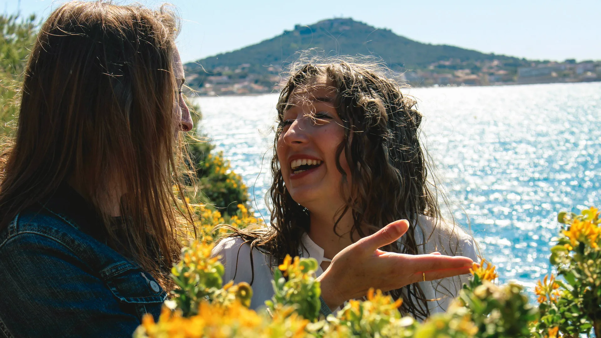 A photograph of two women laughing in conversation in front of yellow flowers against a lake and mountain background