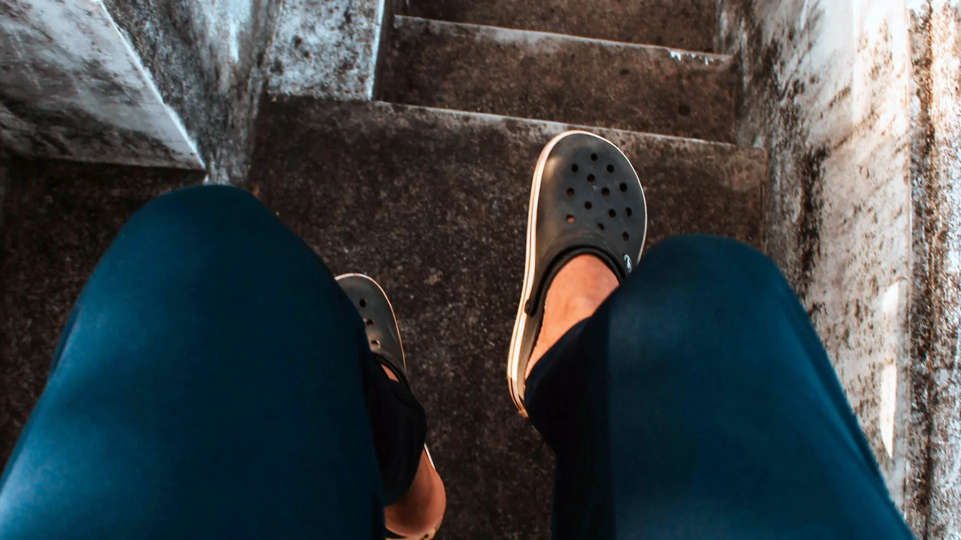 A photograph of a person standing at the top of a stone staircase wearing black croc shoes and blue trousers