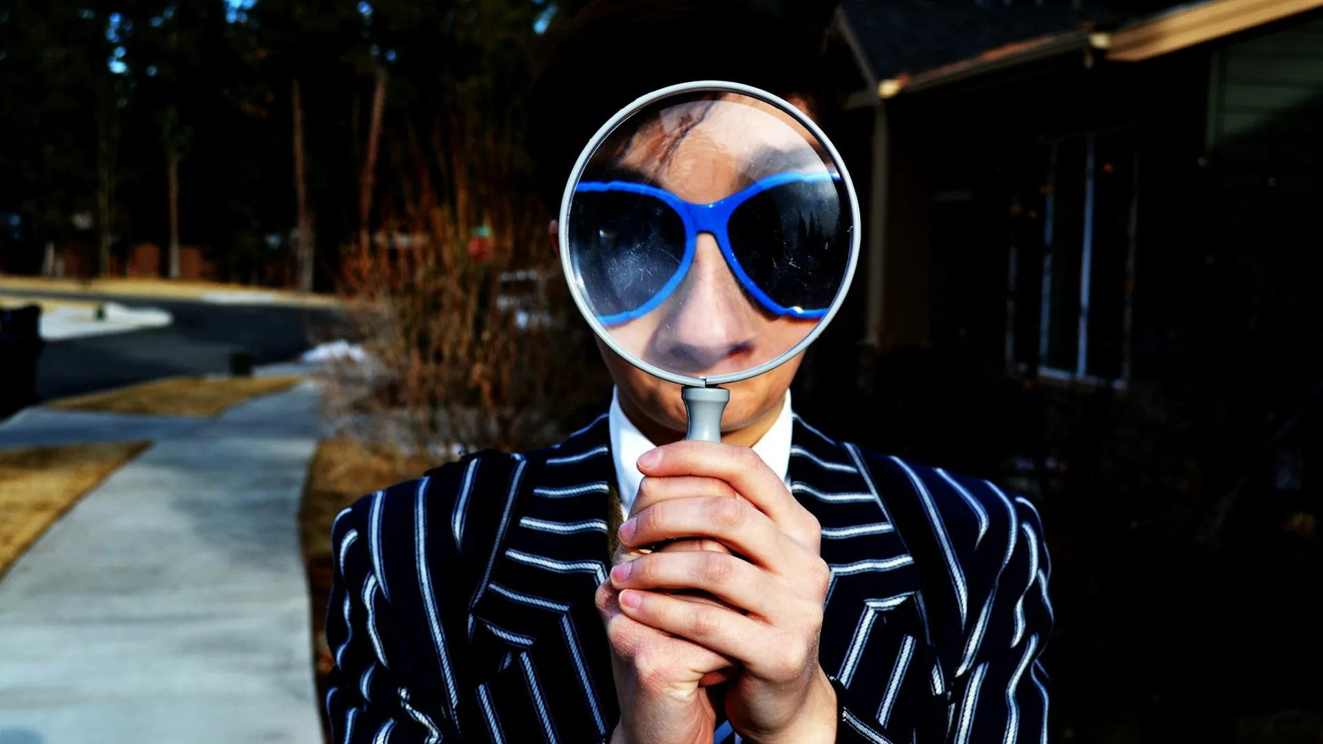Person wearing a pinstriped jacket holds magnifying glass in front of face so it makes their nose and blue glasses very large