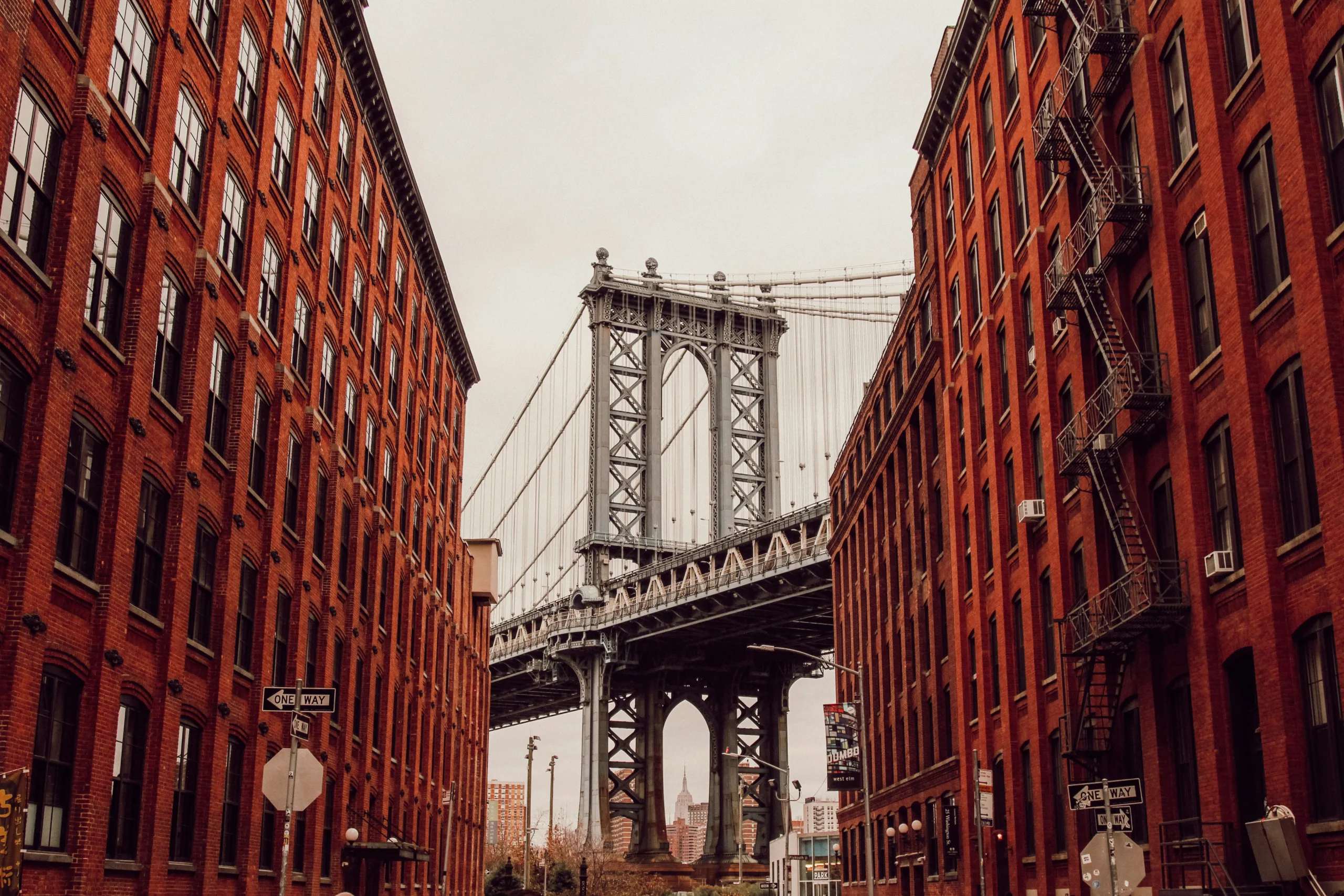A photograph of Brooklyn bridge at the end of a street of red brick buildings against a grey sky