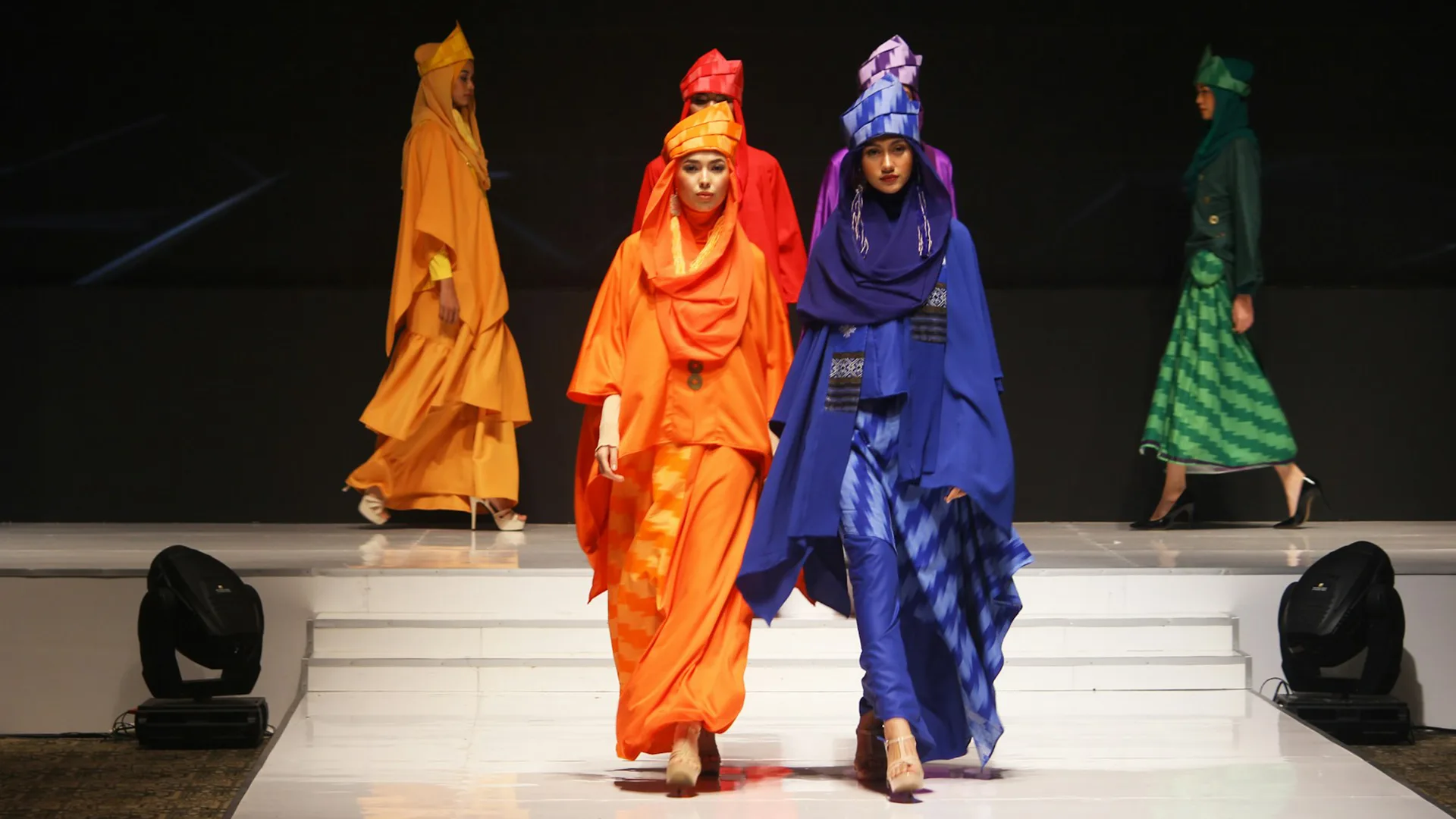 Women walking down a catwalk wearing orange and purple dresses and headscarves