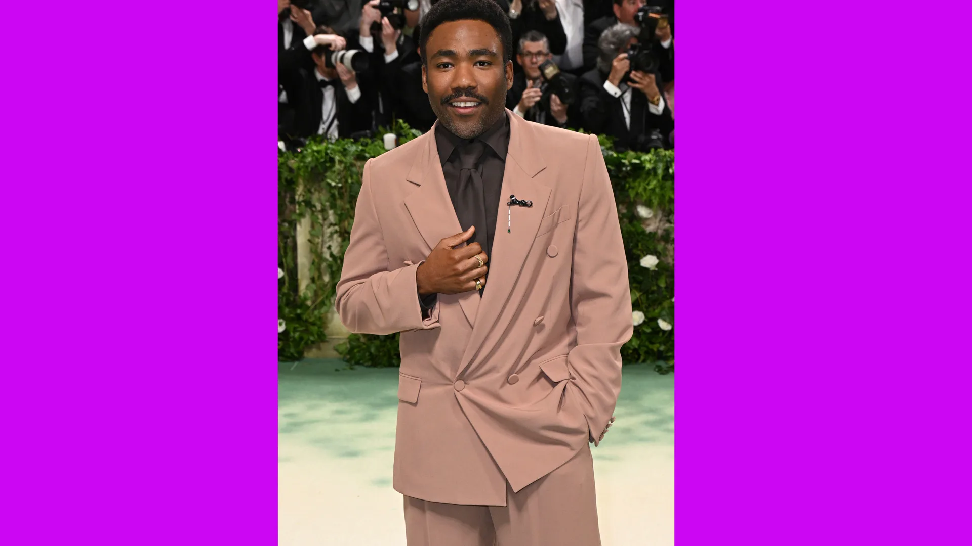 A photograph of Donald Glover at the Met Gala 2024 wearing a brown suit with press photographers behind him