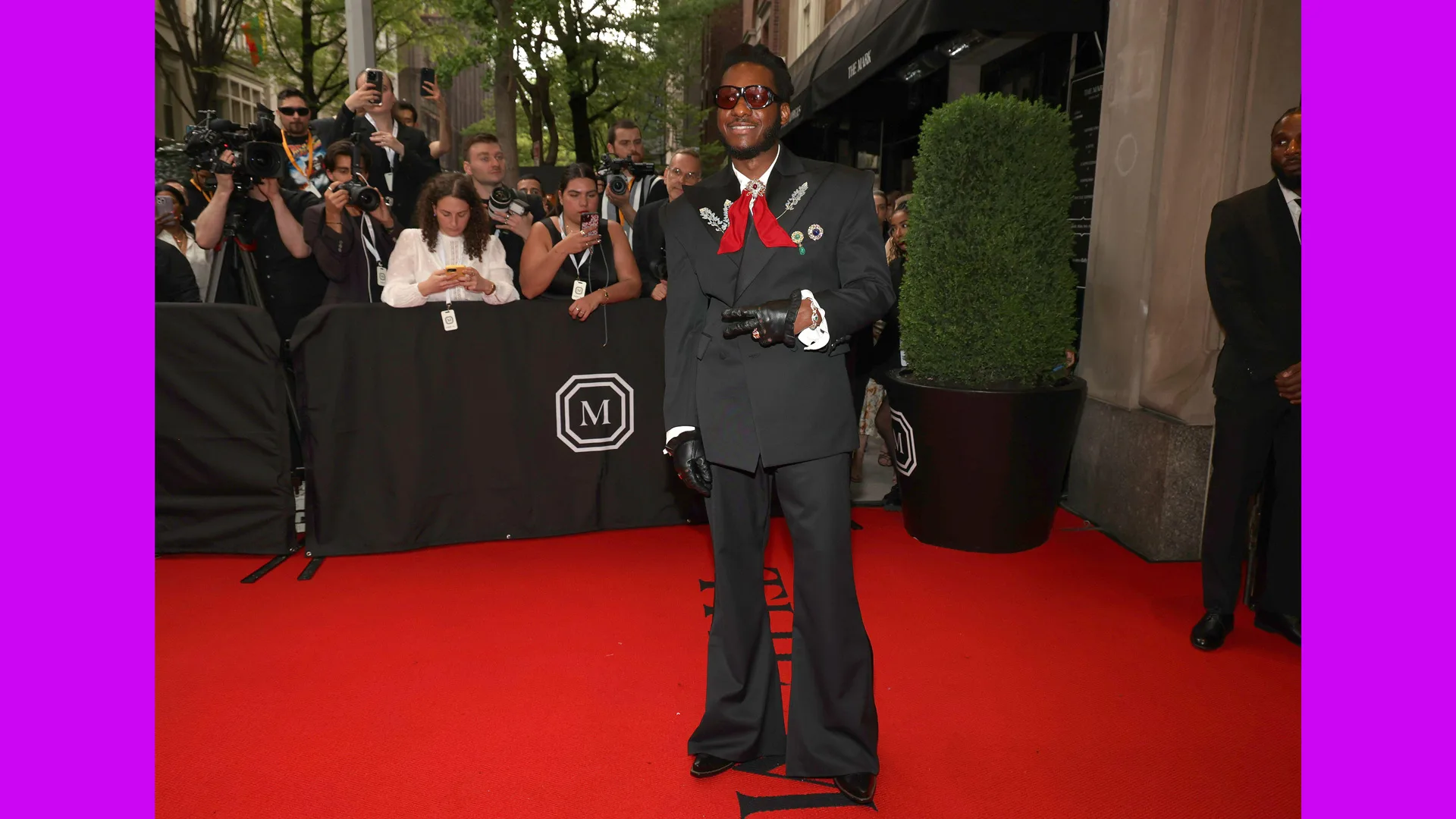 A photo of Leon Bridges on the red carpet at the Met Gala 2024 wearing a black suit with a red bow - press photographers are behind him